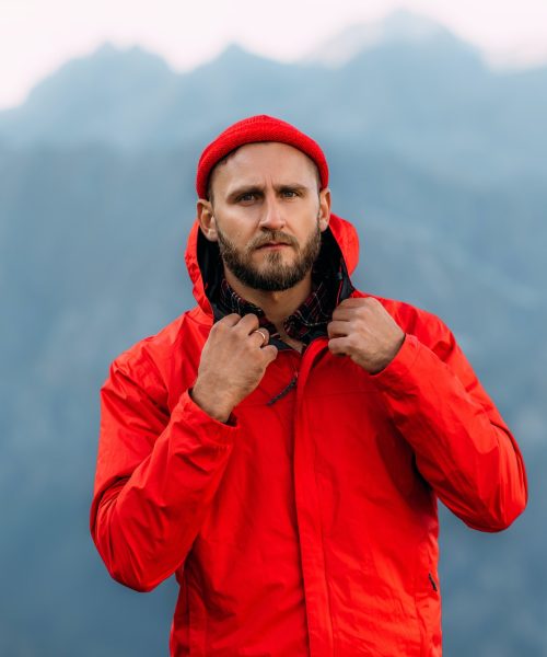 Portrait of a brutal bearded man in a red jacket and hat among the mountains